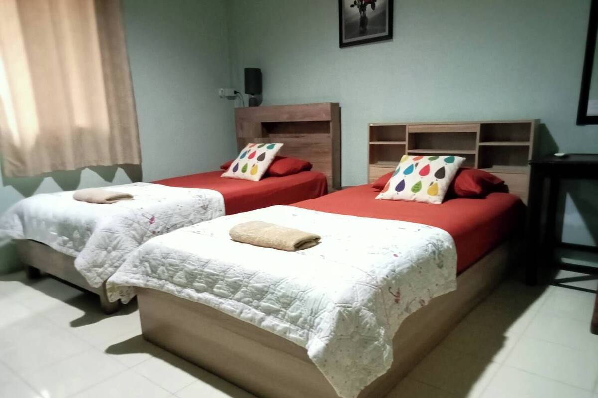 A hotel room with two beds, each with a red mattress and white bedding. There are wooden headboards and a small wooden shelf unit with colorful pillows.