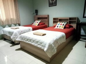 Een hotelkamer met twee bedden, elk met een rode matras en witte beddengoed. Er zijn houten hoofdborden en een klein houten wandrek met kleurrijke kussens.