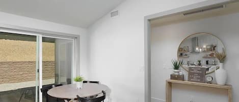 A modern dining area with a round table and chairs, a glass door leading outside, and a decorative shelf with a mirror and plants.