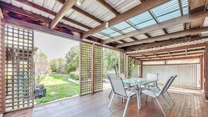 Eine Terrasse im Freien mit einem Glastisch und Stühlen, Holzboden und Blick auf einen Garten.