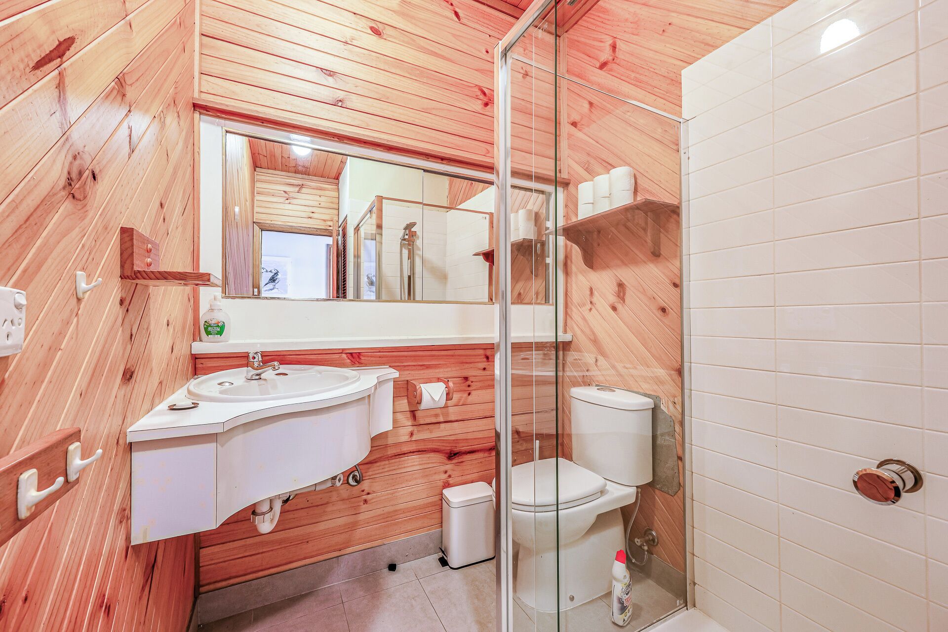 A bathroom with wooden walls, a white sink, a toilet, and a glass shower enclosure.