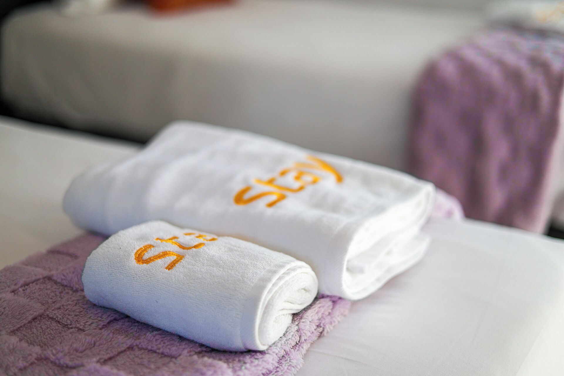 A neatly folded white towel with the text "St. 3" on it, placed on a bed with a purple blanket.