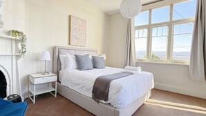 A bedroom with a large bed, a bedside table, a lamp, a window with curtains, and a view of the city. - StayAU 2BR Home w/ Stunning Views in Tasmania (Trevallyn)