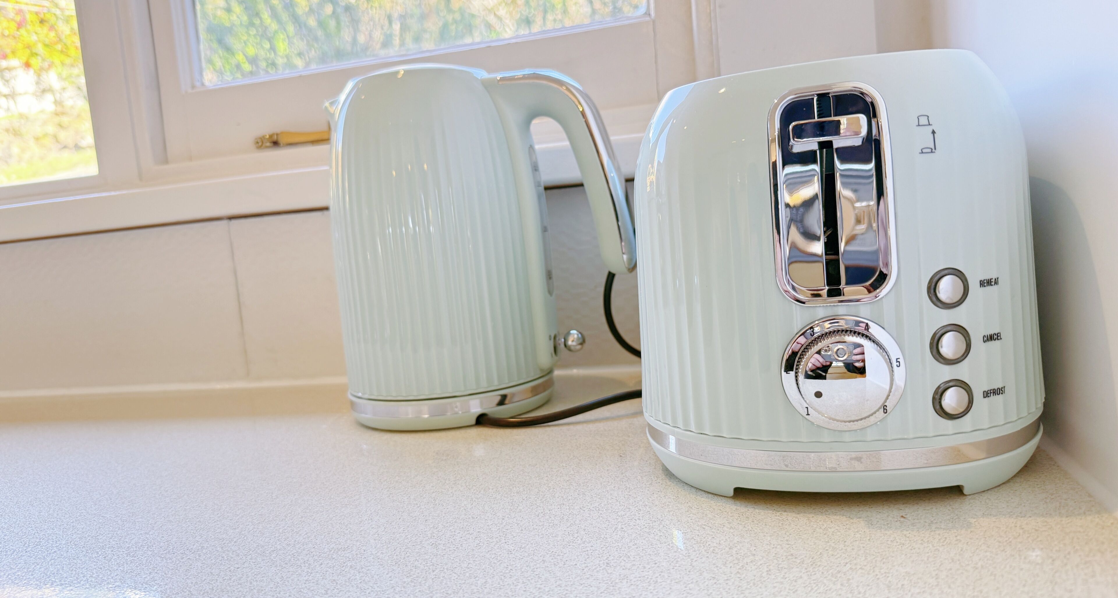 Two kitchen appliances, a kettle and a toaster, on a countertop near a window.