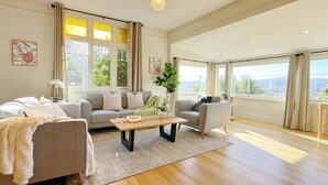 A spacious living room with a grey sofa, a wooden coffee table, and a view of the city through large windows. - StayAU 2BR Home w/ Stunning Views in Tasmania (Trevallyn)