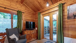 L’intérieur d’un chalet accueillant, avec un plafond en bois, un téléviseur posé sur un meuble en bois, un fauteuil gris, une petite table blanche et une vue sur l’extérieur grâce à une porte vitrée.