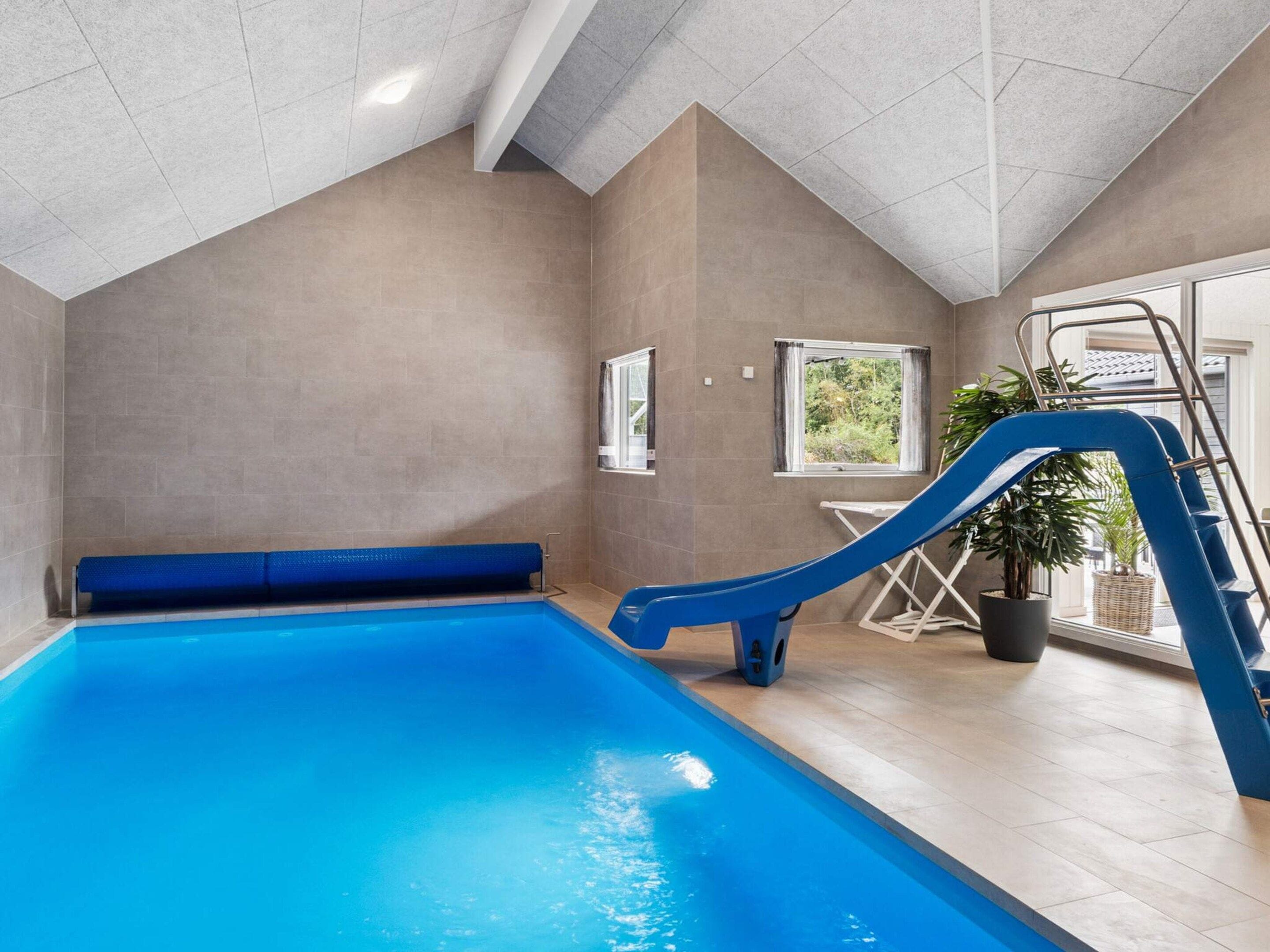 An indoor swimming pool with a blue slide, a window with a view, and a potted plant.