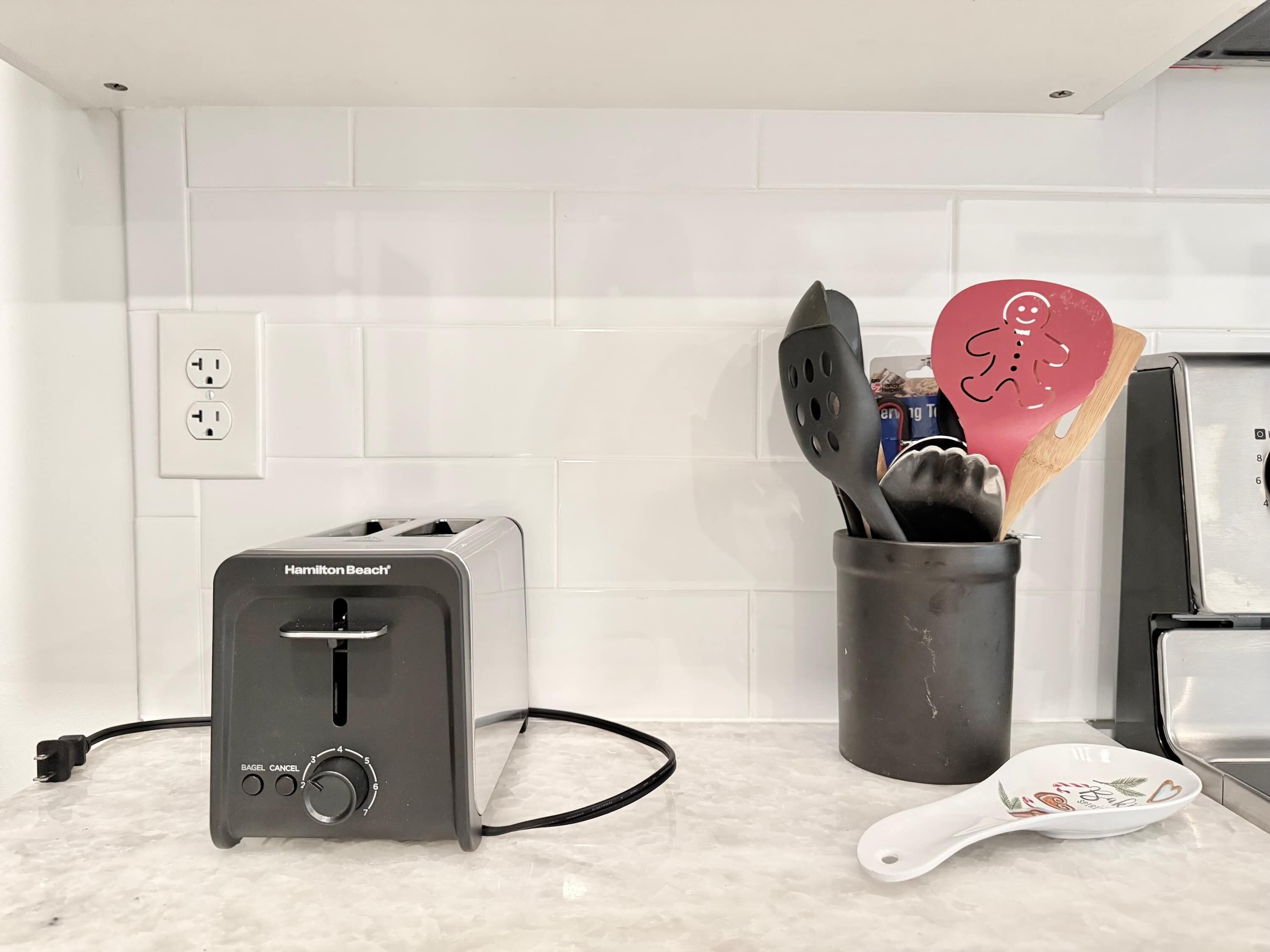 A kitchen with a Hamilton Beach toaster, a utensil holder with spatulas and a wooden spoon, and a white plate with a heart design.