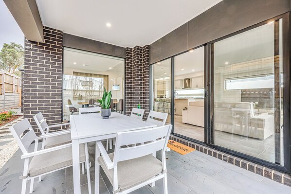 A modern outdoor dining area with a white table and chairs, a potted plant, and a 'WELCOME' mat.