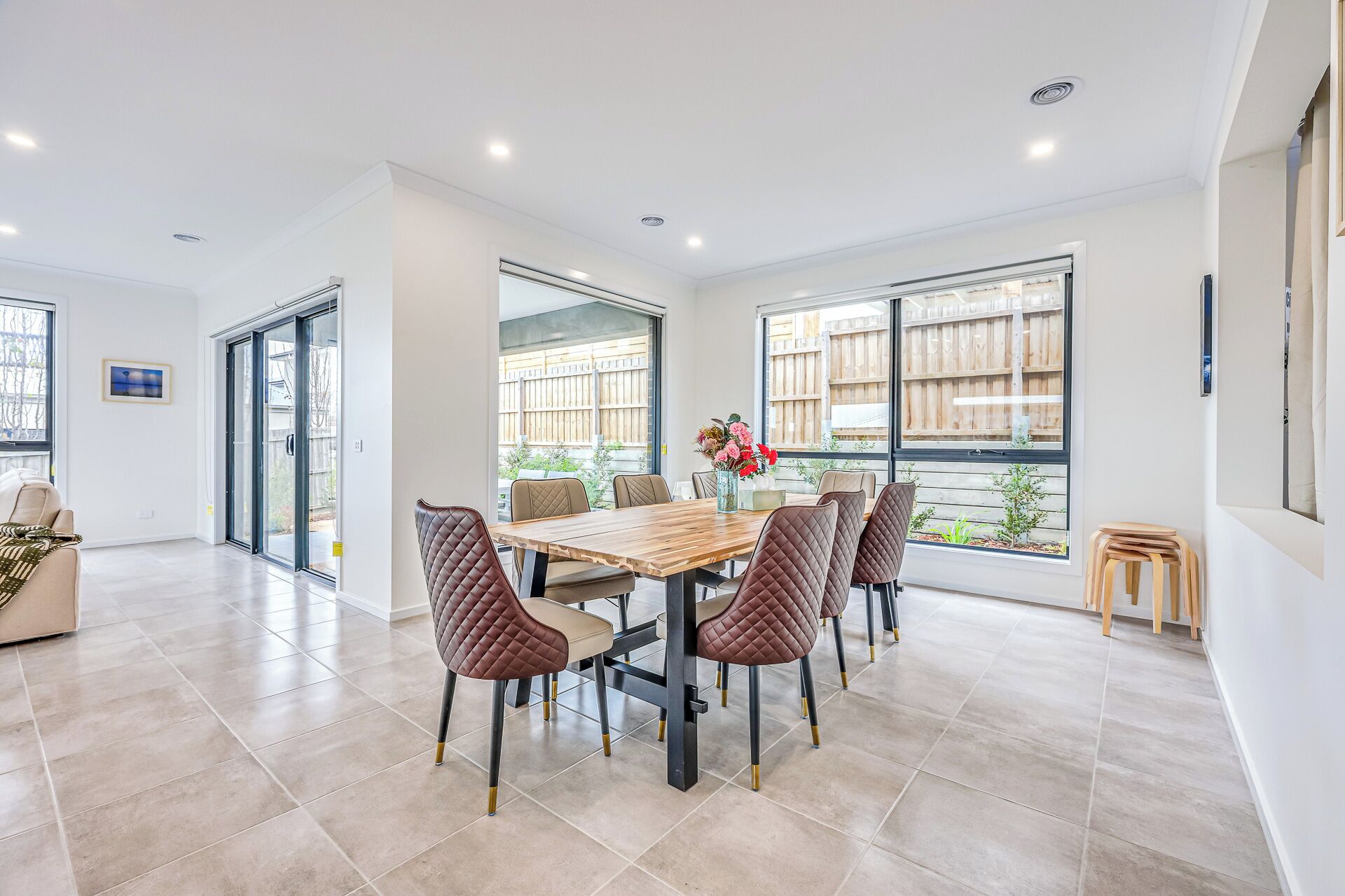 A modern dining area with a wooden table and cushioned chairs, large windows with a view of a wooden fence, and a clean, tiled floor.