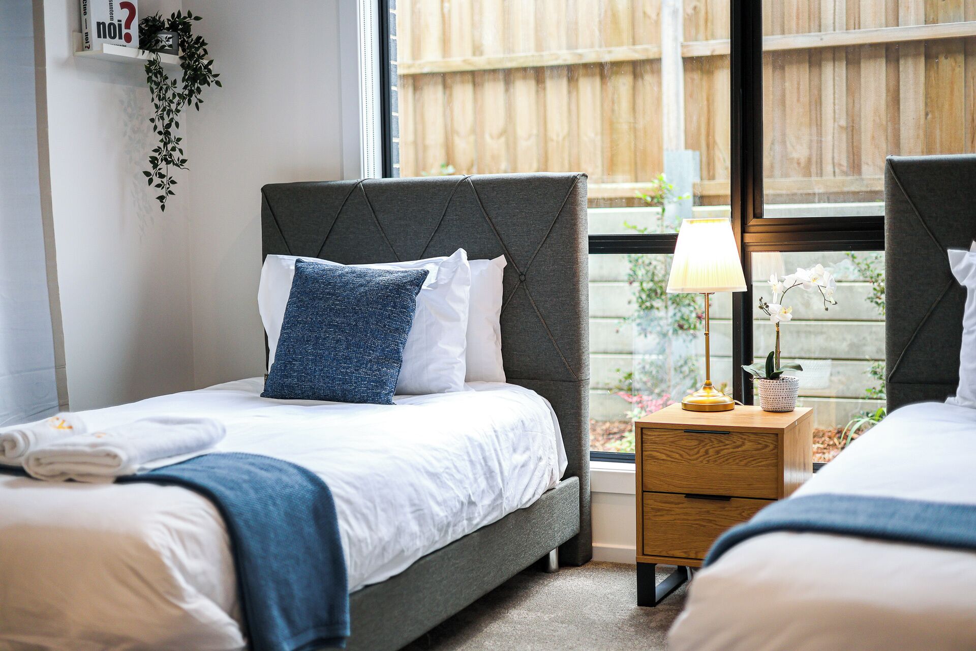A bedroom with two beds, a bedside table with a lamp, and a window with a view of a wooden fence.