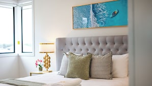 A neatly made bed with a grey tufted headboard, a teal artwork above it, and a window to the left. - StayAU 5BR Family Getaway GreaterGeelong in Duneed (Mount Duneed)