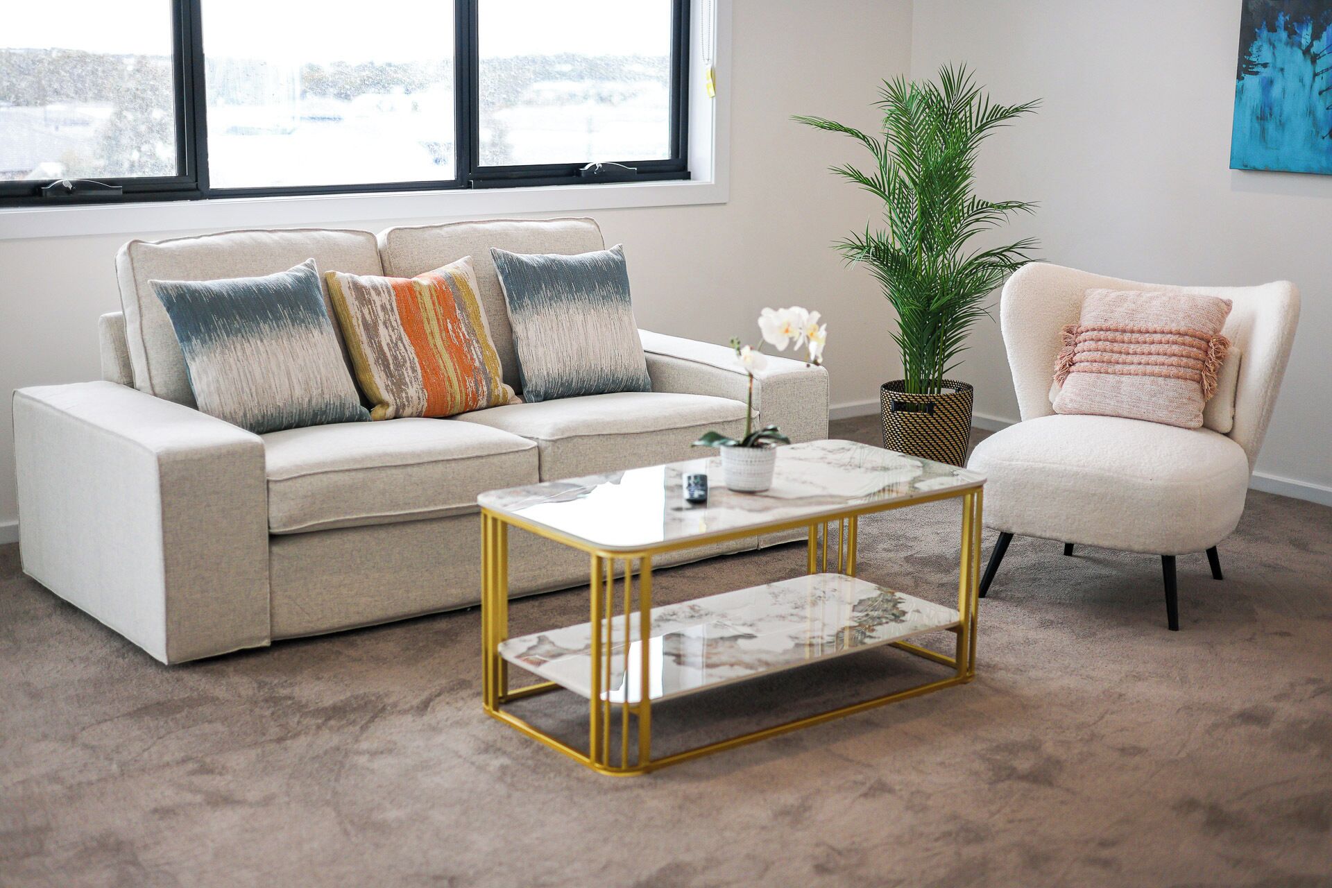 A living room with a beige sofa, a white armchair, a glass coffee table, and a potted plant.