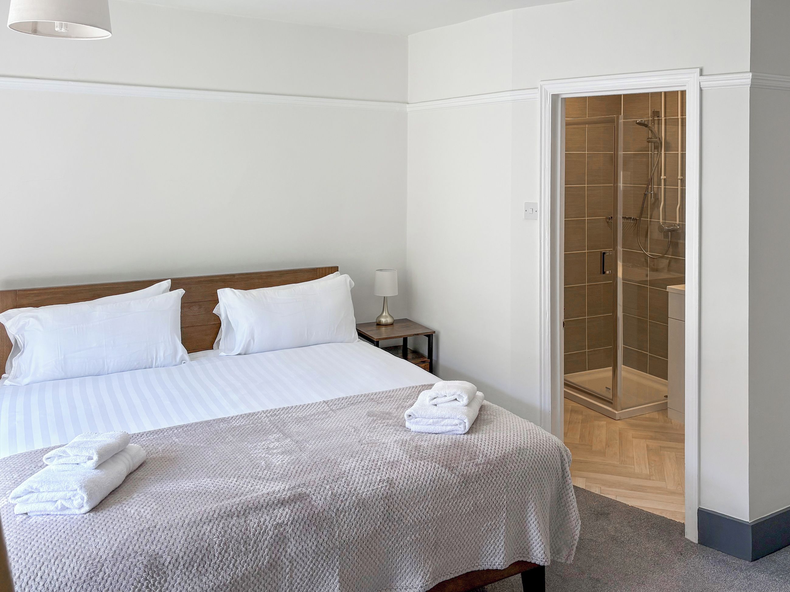 Un lit bien fait, avec du linge de lit blanc et une serviette pliée, une table de chevet avec une lampe, et une douche à l’italienne avec une porte en verre.