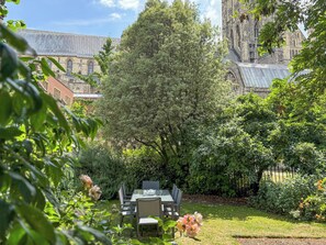 Un jardin avec une table et des chaises, entouré d’une végétation luxuriante et avec un bâtiment historique en arrière-plan.