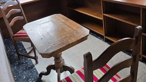 Een zithoek met een houten tafel en stoelen, grote ramen met uitzicht op bomen, en een vloerbedekking.