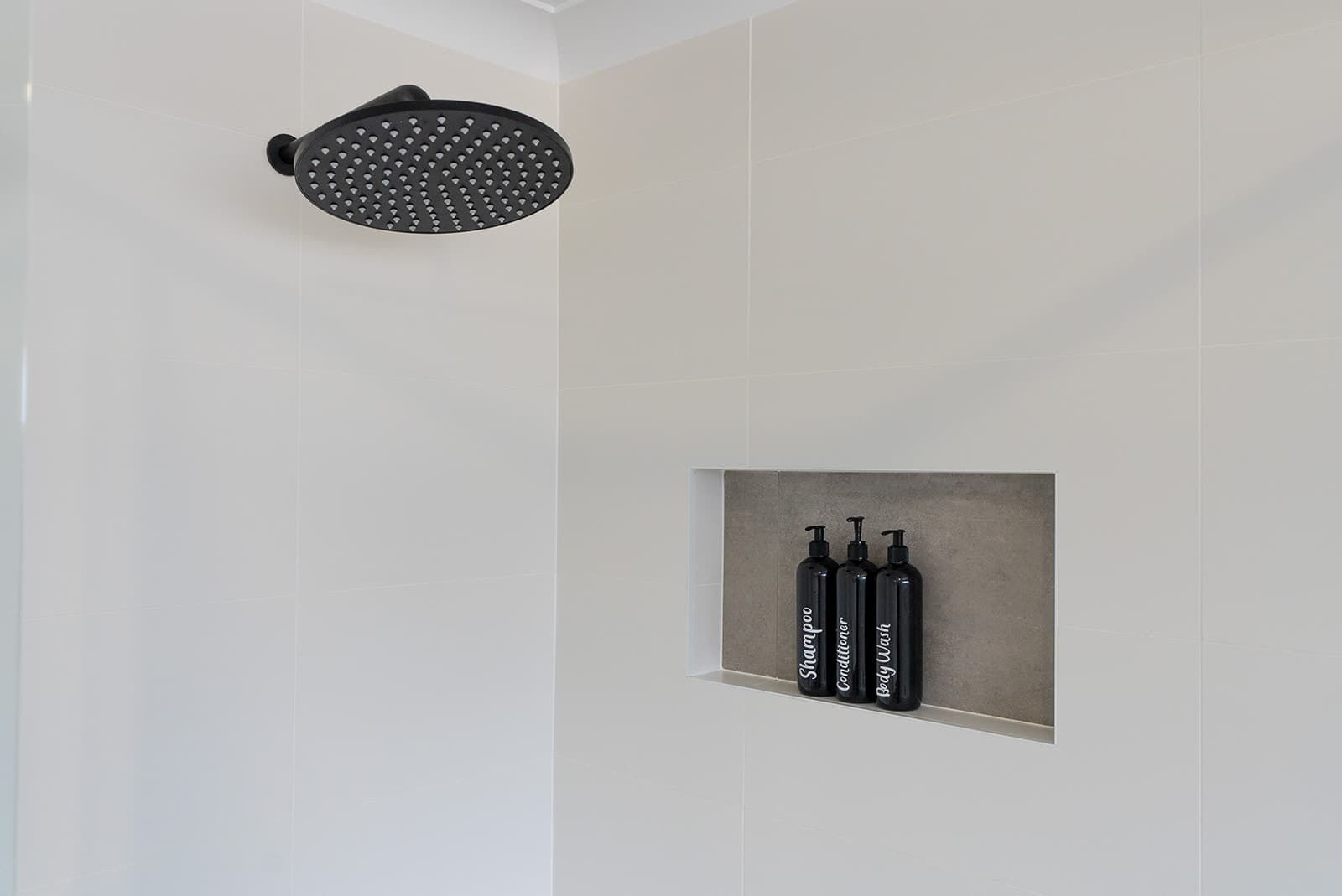 Ducha con cabezal redondo y estante con tres botellas negras etiquetadas champú, acondicionador y gel de baño.