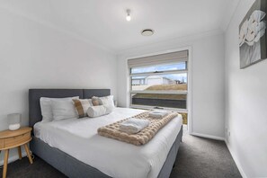A modern bedroom with a large bed, a bedside table, a window with a view of a building, and a ceiling light. - Ignatius Retreat | Bathurst | Family Entertainer (Kelso)