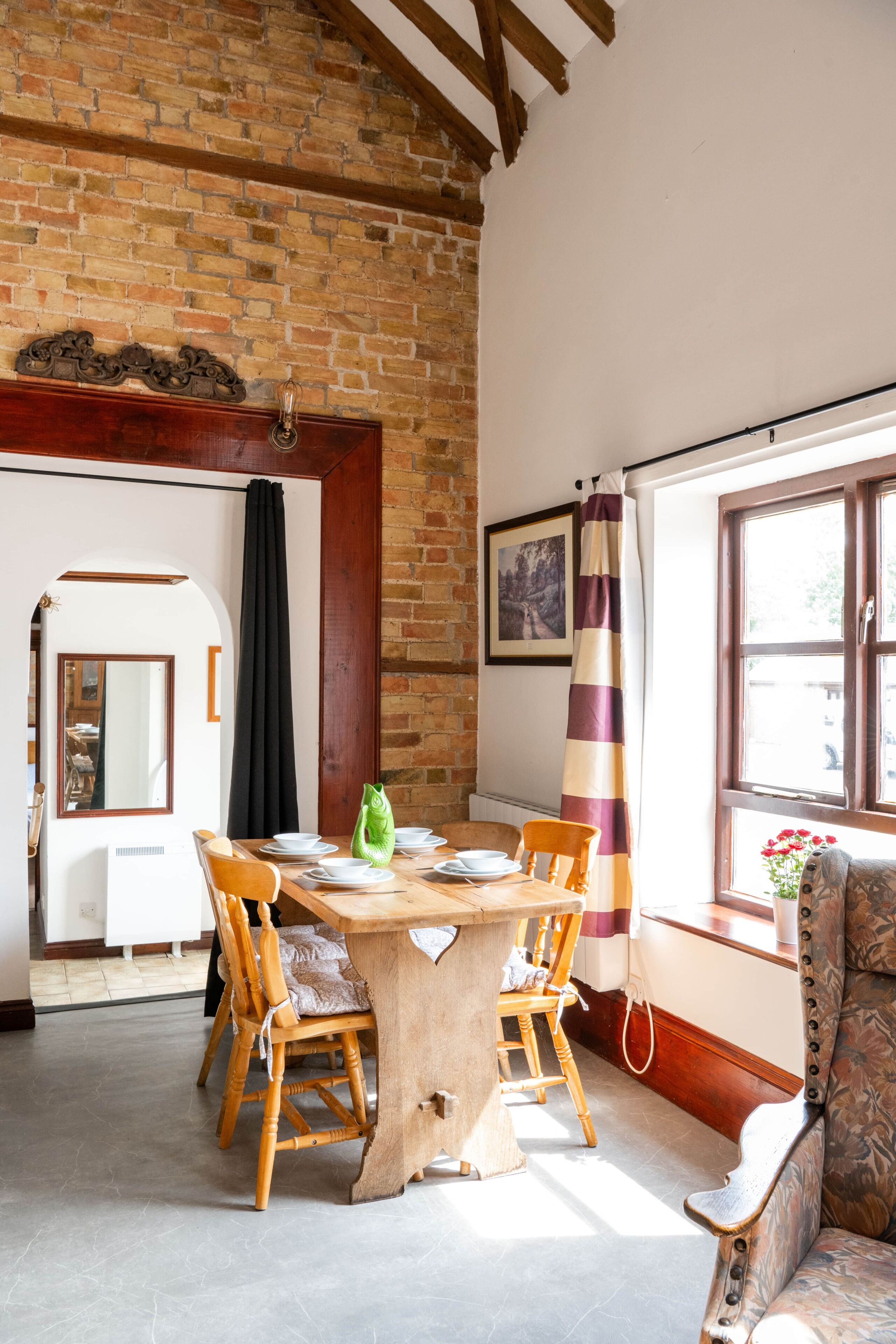 A dining area with a brick wall, wooden ceiling beams, a table with chairs, and a window with curtains.
