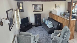 A cozy living room with a television, sofa, and chairs, wooden beams on the ceiling, and a kitchen area with a sink and cabinets.