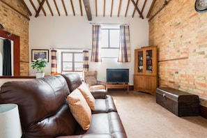 A living room with a leather sofa, a television, a cabinet with glass doors, and a brick wall. - Cottage 2 at Scald end farm (Bedford)