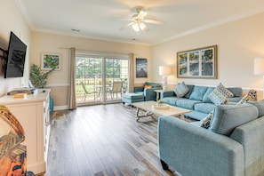 A spacious living room with a large sofa, a coffee table, and a ceiling fan. There is a sliding glass door leading to an outdoor patio area.