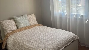 A neatly made bed with a patterned bedspread, a hanging lamp, sheer curtains, and a view of buildings outside.