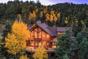 Une cabane en rondins dotée d’une grande façade vitrée, entourée d’un feuillage automnal et située au pied d’une colline boisée.