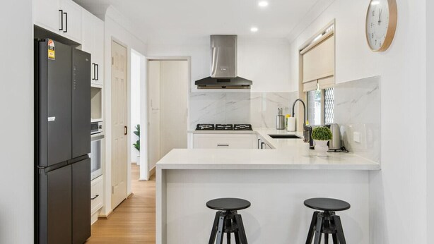 A modern kitchen with white cabinets, a black refrigerator, a stainless steel range hood, a marble countertop, and two black stools. - Spacious Family Retreat in Peaceful Parkinson (Parkinson)