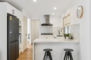 A modern kitchen with white cabinets, a black refrigerator, a stainless steel range hood, a marble countertop, and two black stools. - Spacious Family Retreat in Peaceful Parkinson (Parkinson)