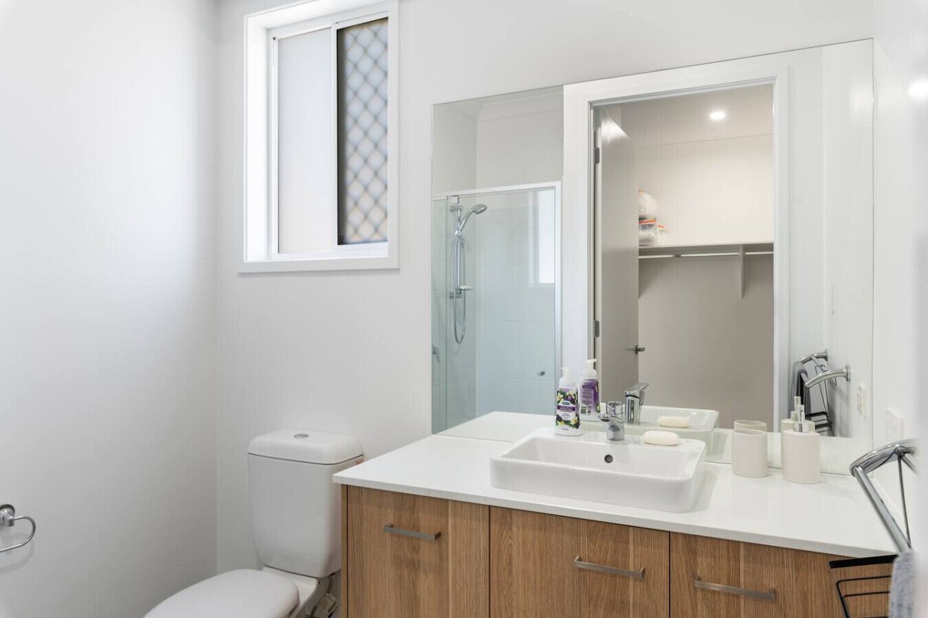 Un baño moderno con lavamanos blanco, armarios de madera, cabina de ducha de vidrio y una ventana sobre el inodoro.