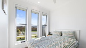 A bedroom with a large bed, three windows, and a view of a residential area.