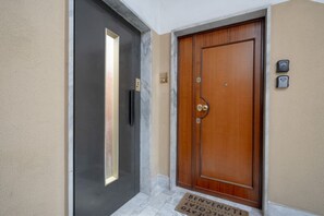 A hotel entrance with a wooden door and a black door, a doormat, and a wall-mounted intercom.