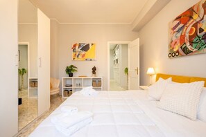 A hotel room with a bed, a bedside table, a lamp, a mirror, a bookshelf, and a painting on the wall.