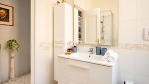 Une salle de bain avec un meuble-lavabo blanc, un miroir, une douche et une plante en pot.