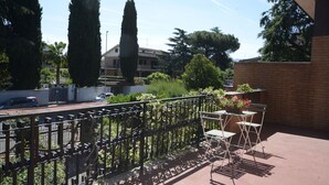 Un balcon avec une balustrade en métal, une table et des chaises. On y trouve des plantes en pot et une vue sur un quartier résidentiel avec des maisons et des arbres.