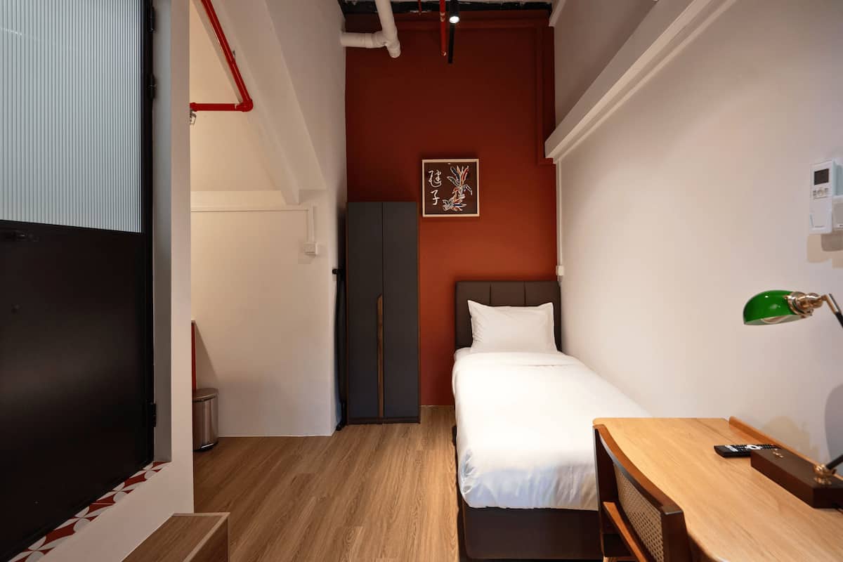 A compact hotel room with a single bed, a desk with a chair, a small table, a green desk lamp, and a wall-mounted air conditioning unit.