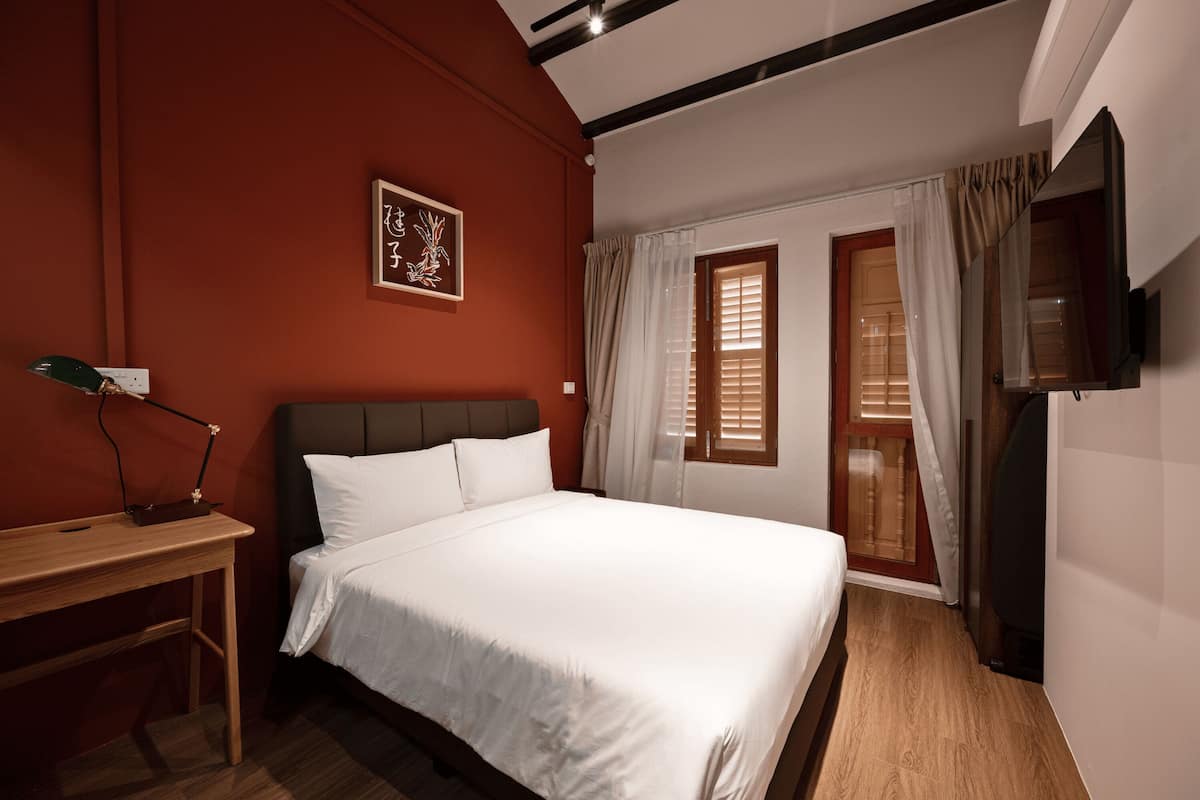 A bedroom with a bed, a desk, a chair, a TV, and a window with curtains.