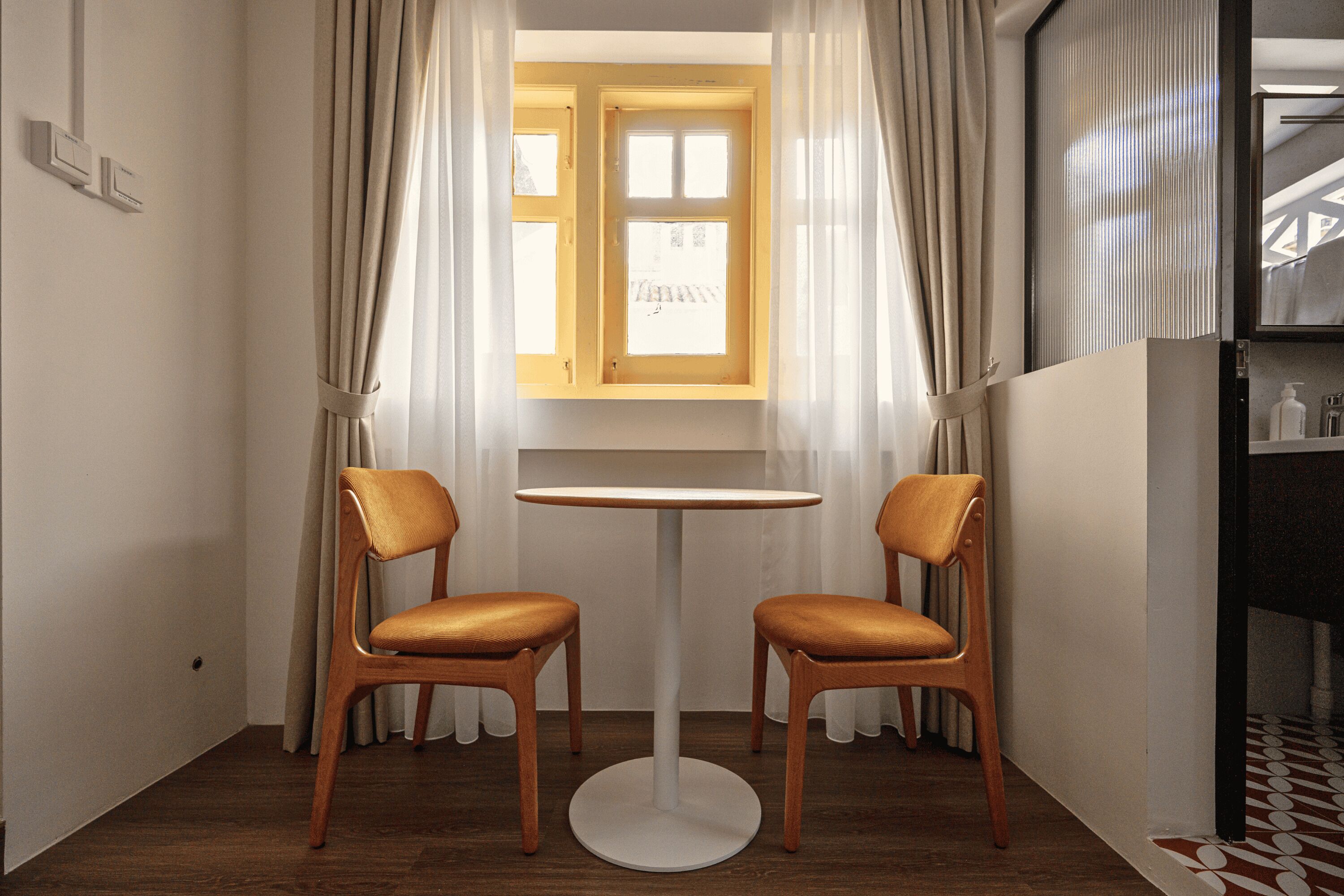 A small room with a round table and two chairs, a window with curtains, and a tiled floor.