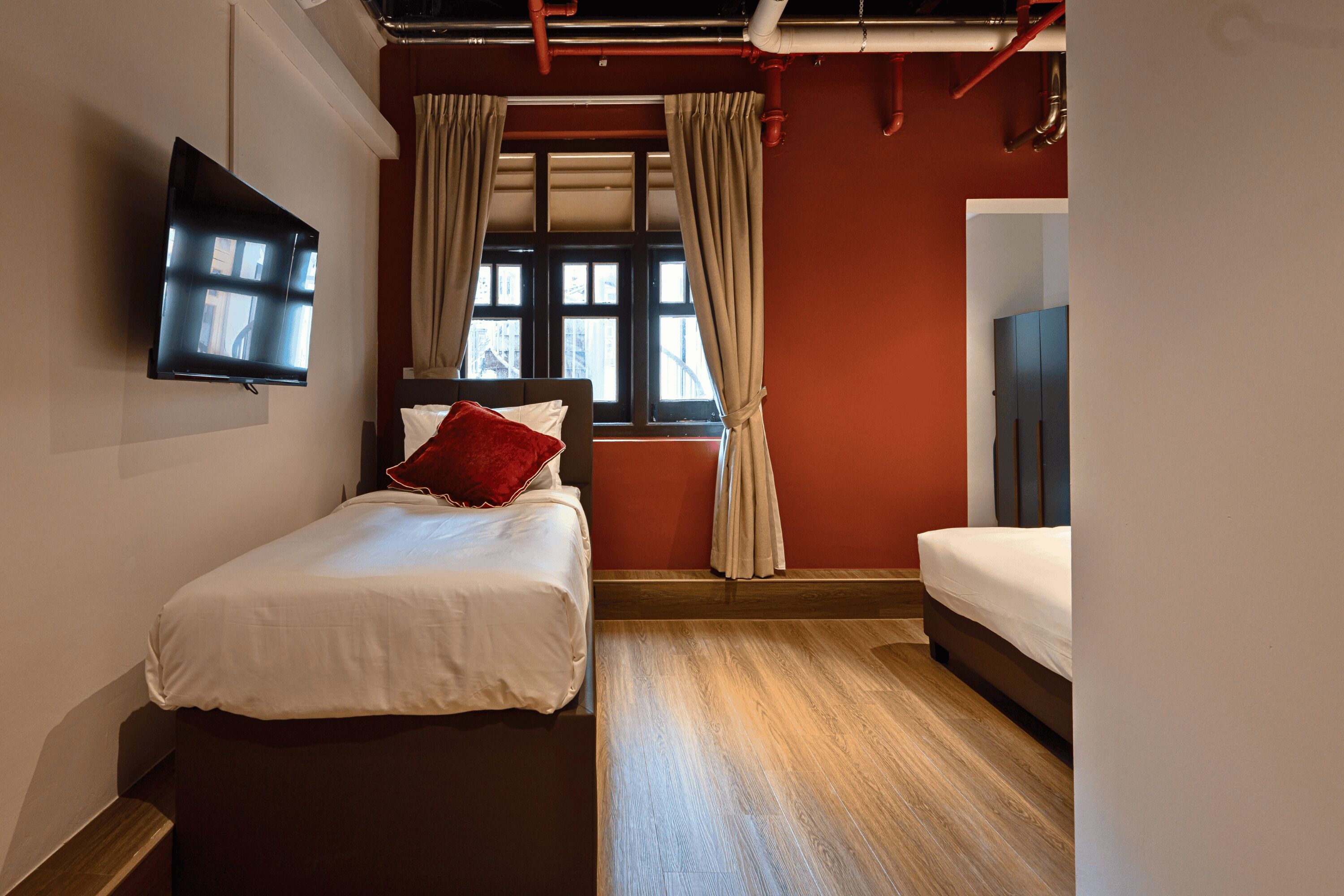 Habitación de hotel con cama, televisión montada en la pared, ventana con cortinas y piso de madera.