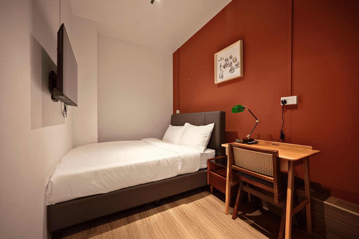 A bedroom with a bed, bedside table, desk, chair, lamp, and a wall-mounted TV.