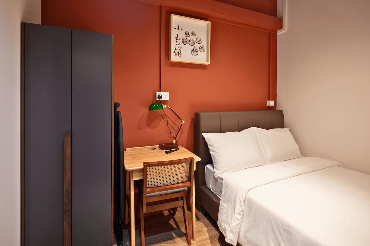 A hotel room with a bed, a desk with a chair, a wardrobe, and a wall-mounted lamp.