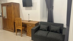Ein Zimmer mit Holzschrank, Schreibtisch und Stuhl, grauem Sofa, flachbildfernseher an der Wand und Fenster mit grauen Vorhängen.