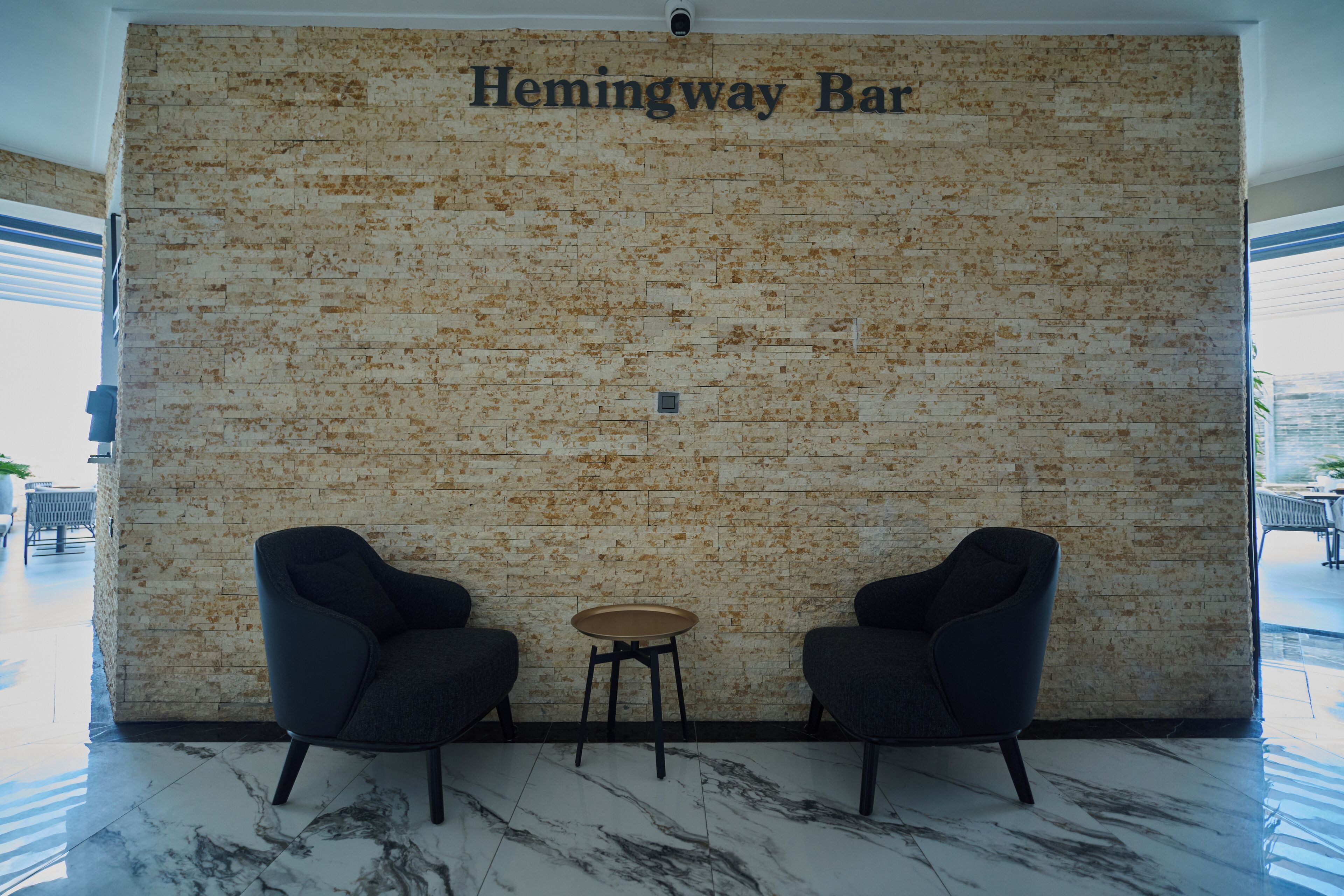 A seating area with two chairs and a small table, a brick wall with "Hemingway Bar" text, and a view of the outdoors.