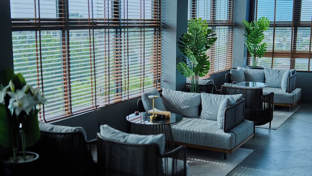 Una sala de estar moderna con sofá, sillones y una mesa de centro. Amplias ventanas con persianas dejan entrar luz natural.
