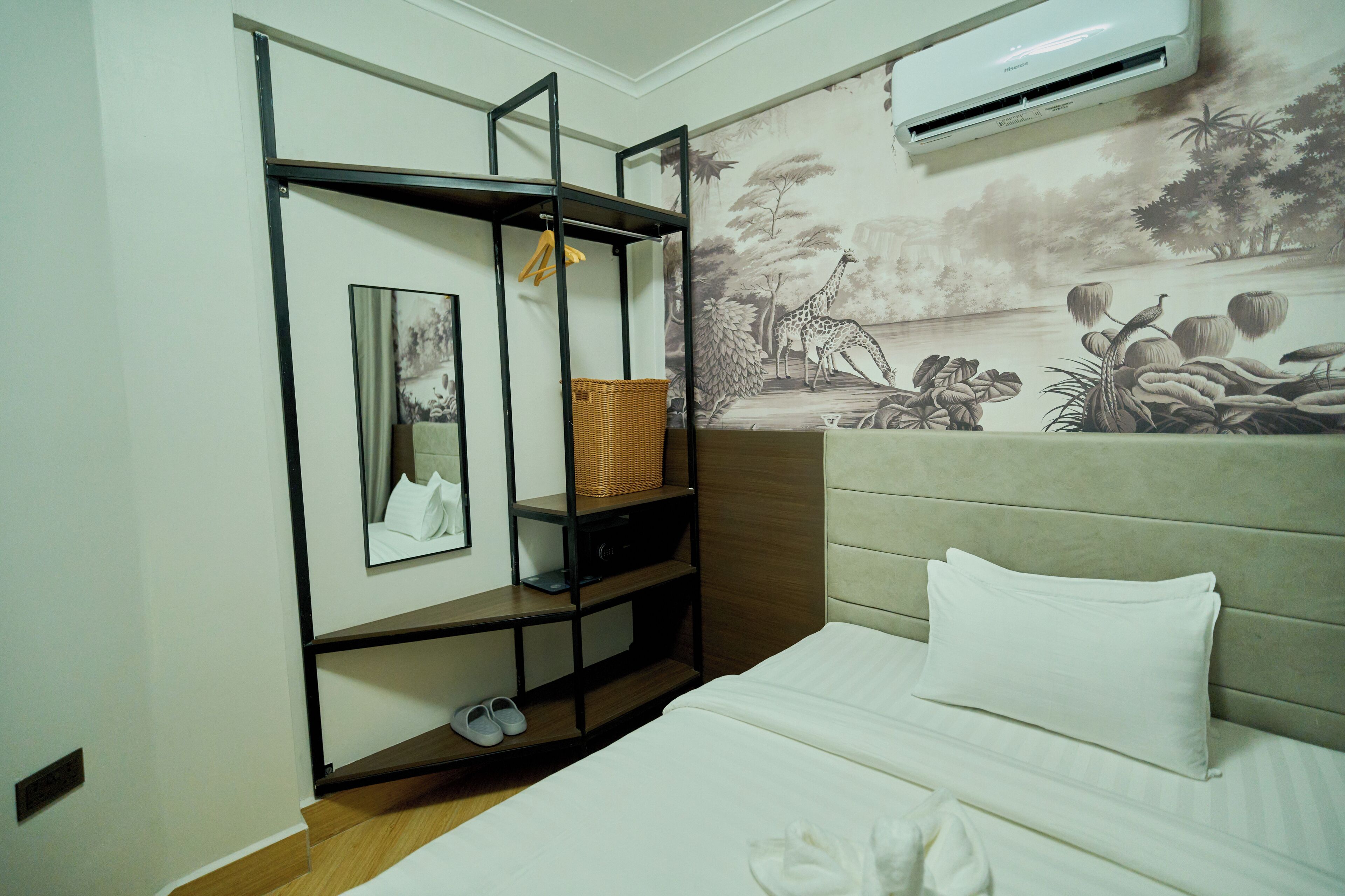 Une chambre d’hôtel avec un lit, une étagère fixée au mur avec un panier, un tableau encadré et une fresque représentant un paysage.
