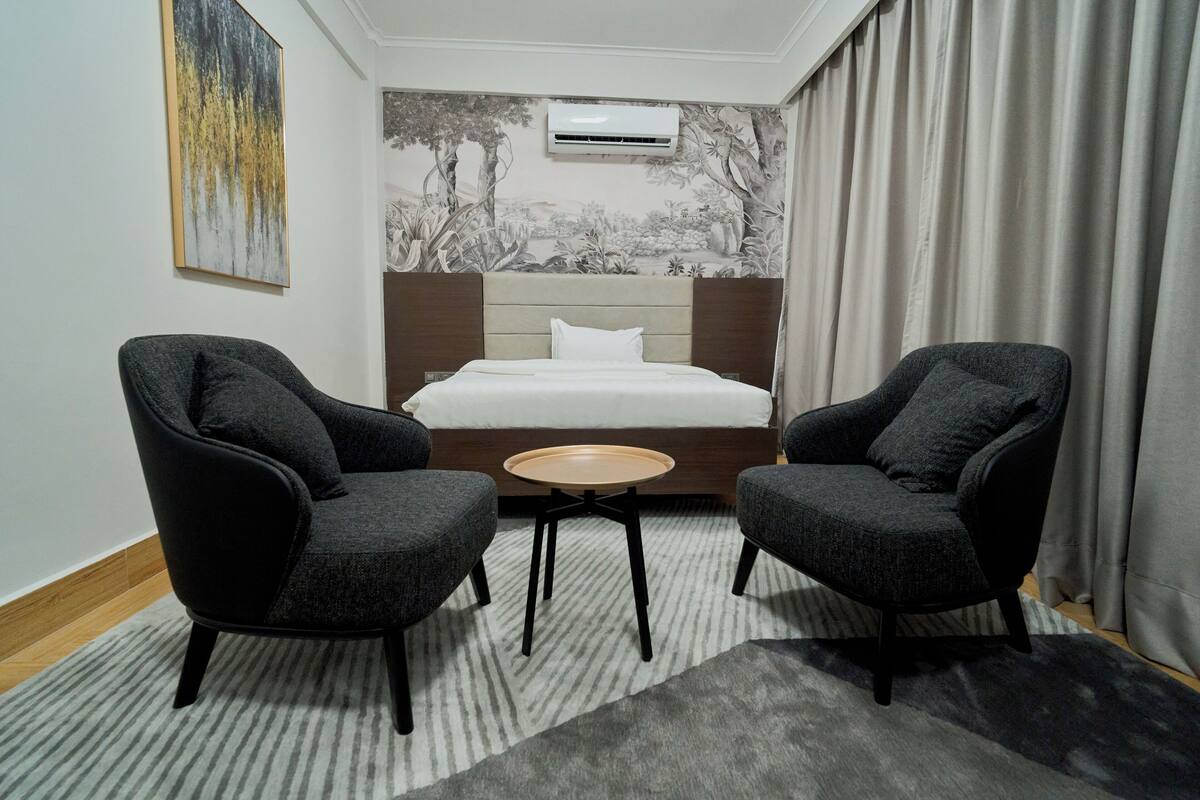 Een hotelkamer met een bed, twee fauteuils, een klein rond tafeltje en een wandairconditioner.