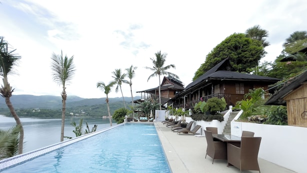An infinity pool with a view of the sea and mountains, lounge chairs, and a wooden building with a thatched roof. - Villa De Pico Highland Beach Resort (Puerto Galera)