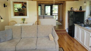 A cozy living room with a grey sofa, wooden ceiling beams, a dining area with a table and chairs, and a kitchen with white cabinets and a black refrigerator. - Whimsical Wildwood Studio – Breakfast Included – Award-Winning Appalachian B&B (Irvine)