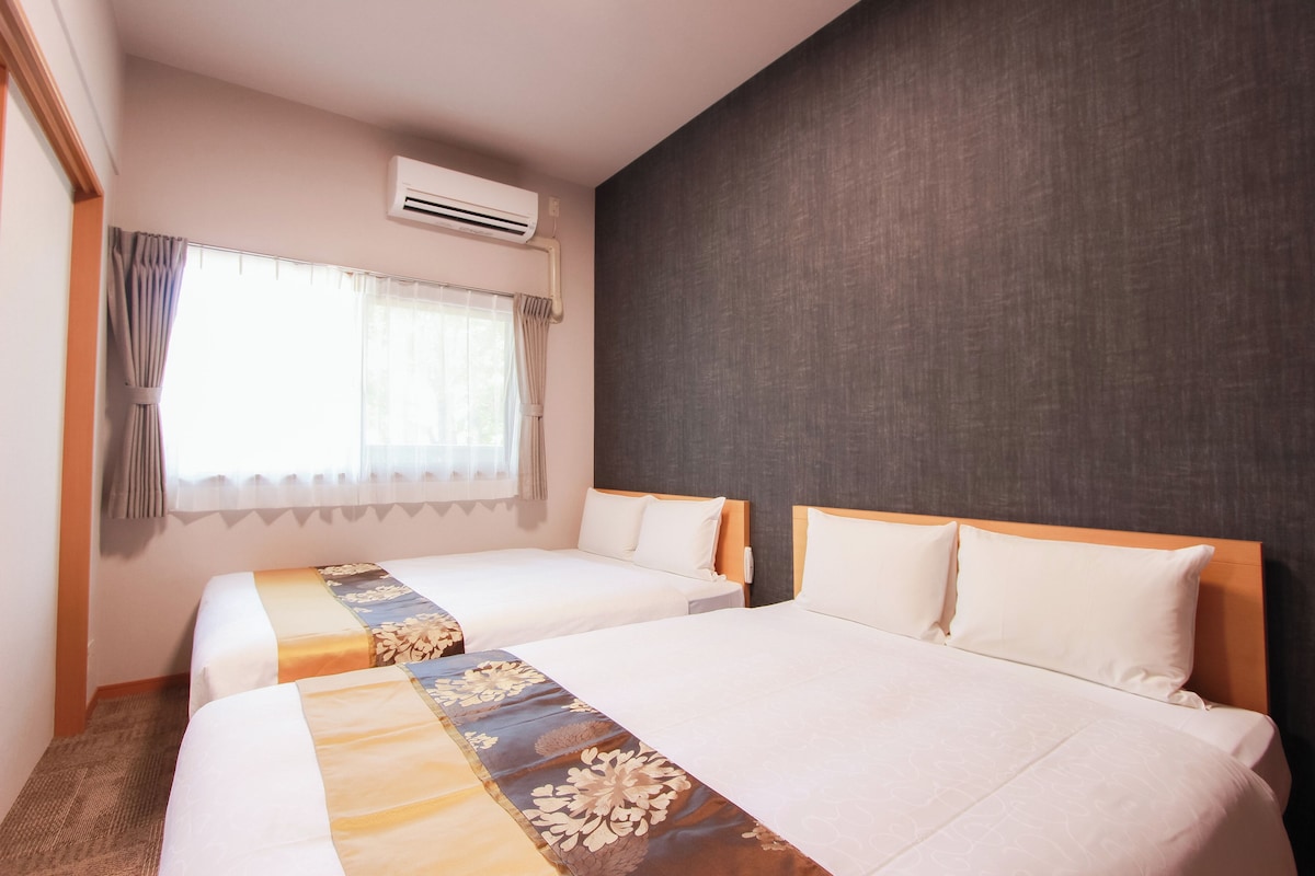Una habitación de hotel con dos camas, una ventana con cortinas, un equipo de aire acondicionado y una pared lisa.